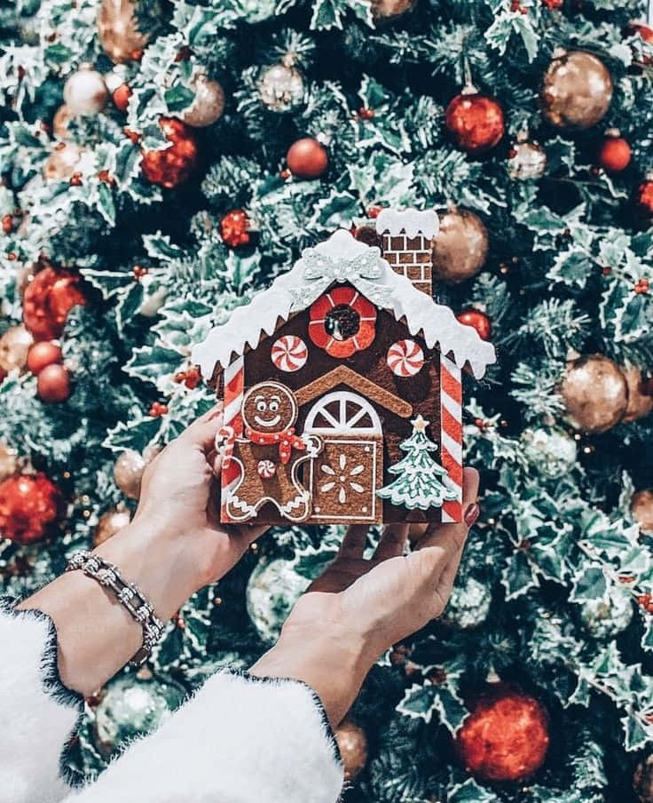 mtu anayeshikilia nyumba ya gingerbread mbele ya mti wa krismasi Picha ya Ukuta